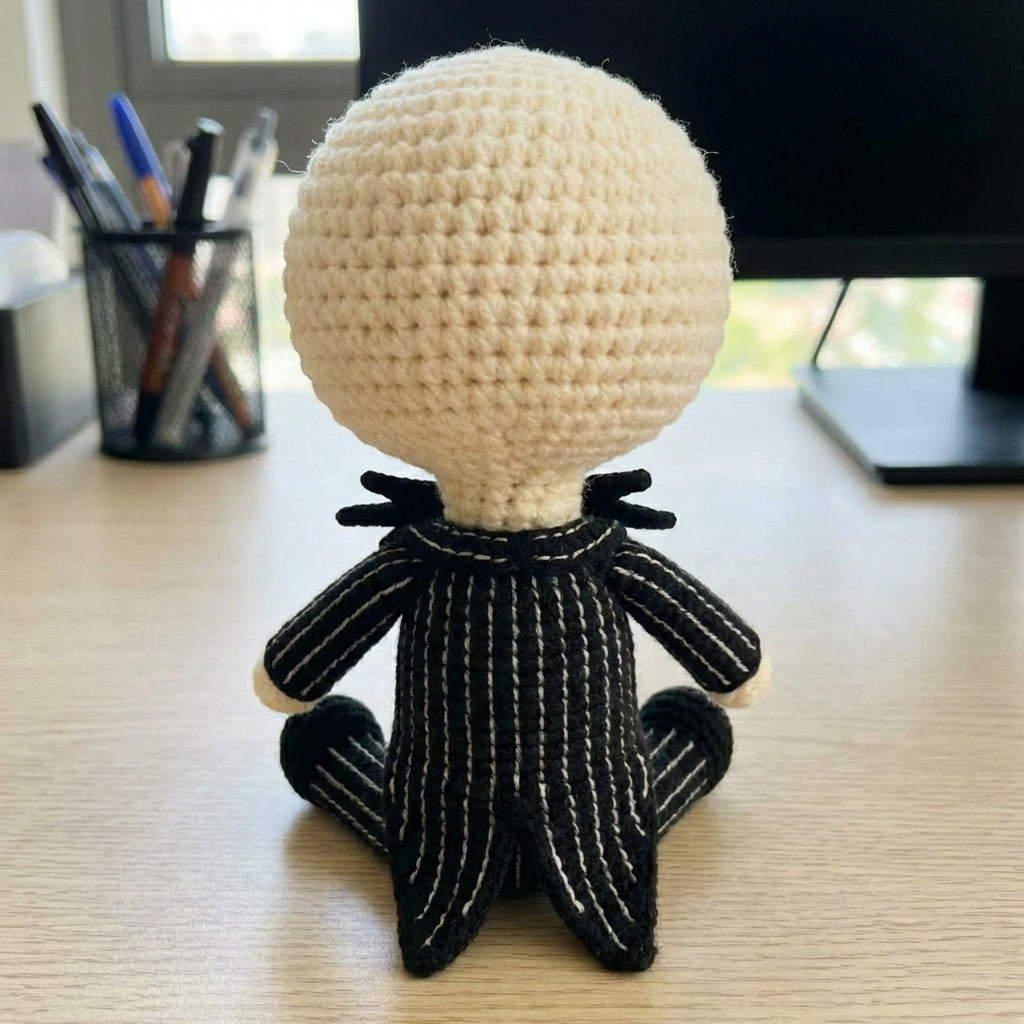 Crocheted character with a round beige head and black body on a desk.