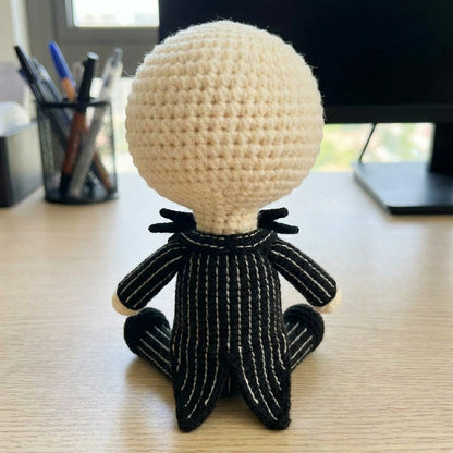 Crocheted character with a round beige head and black body on a desk.