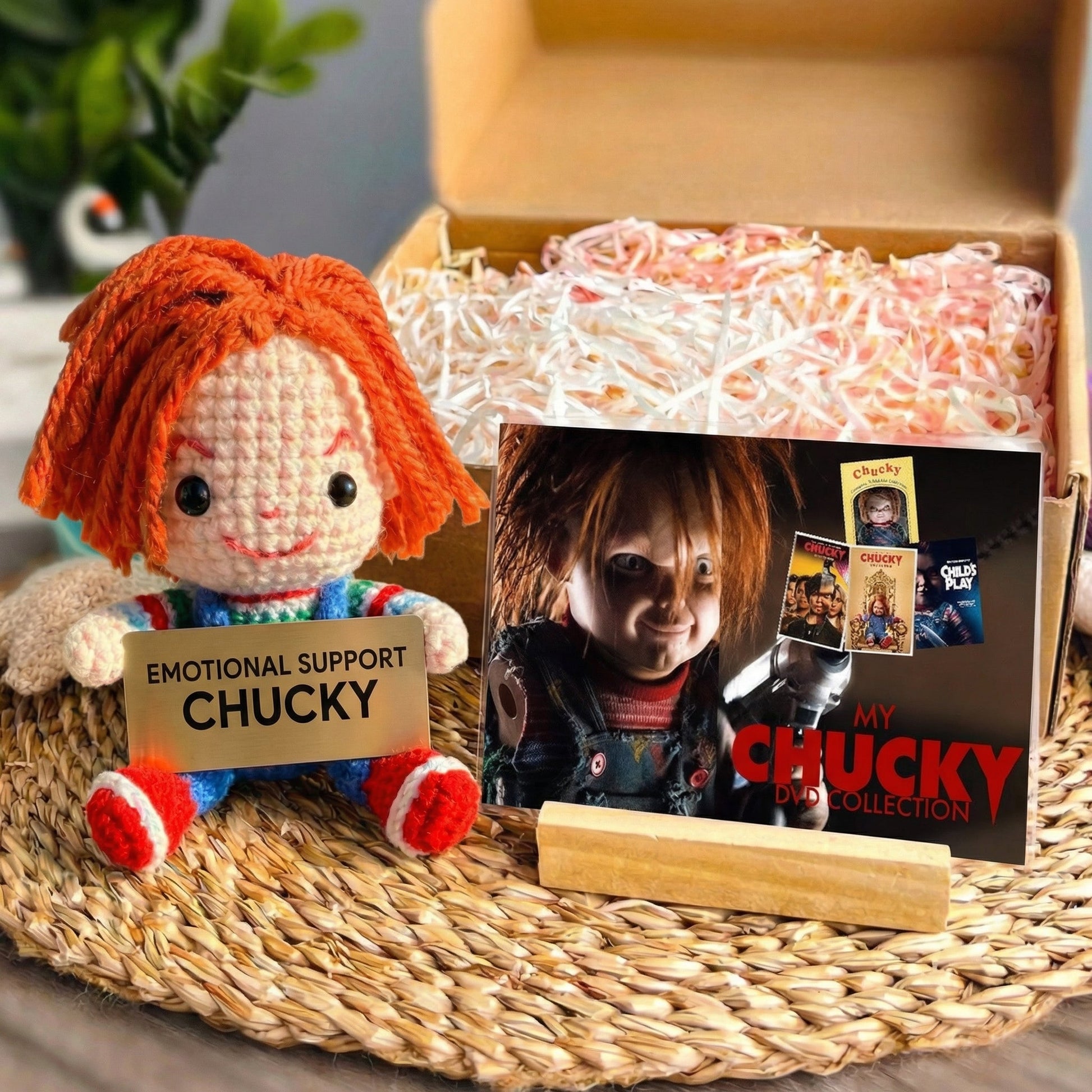 Emotional Support Chucky doll with a box of candy and 'My Chucky Doll Collection' book on a woven surface.