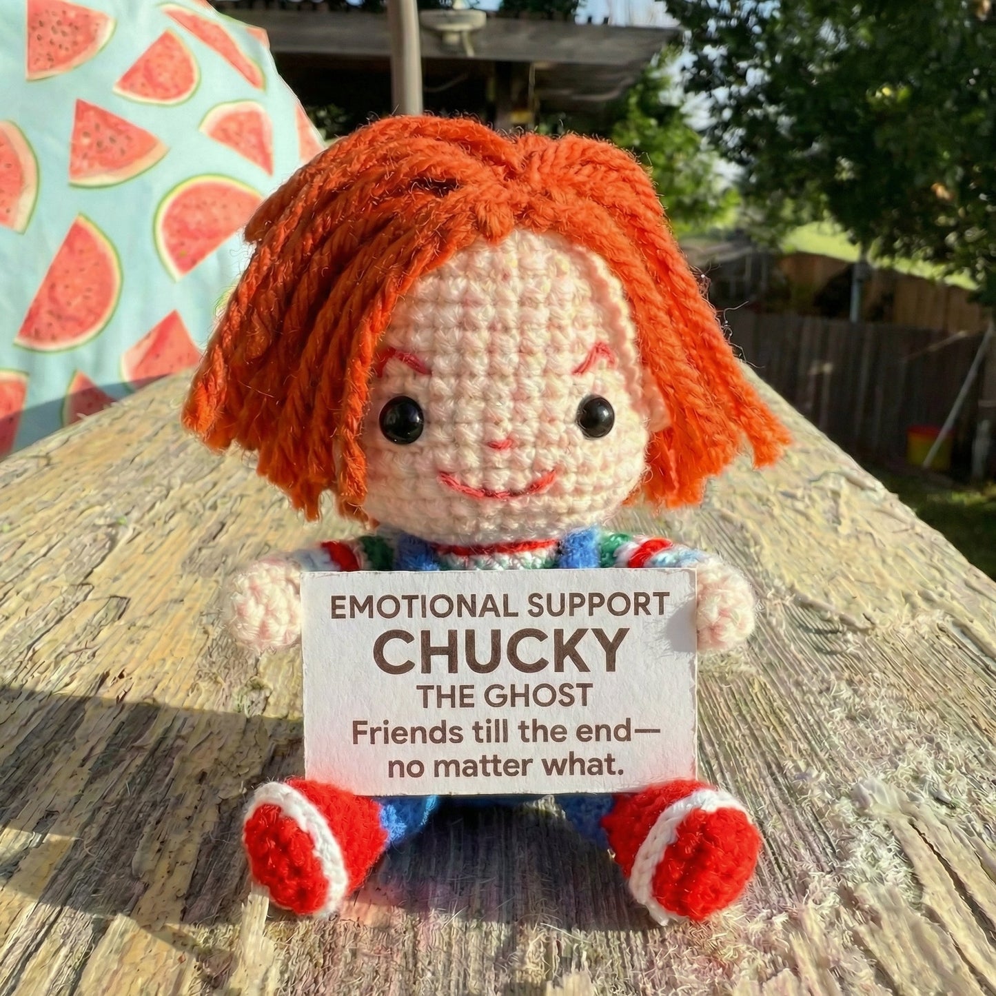 Crocheted doll with orange hair holding a sign on a wooden surface outdoors.